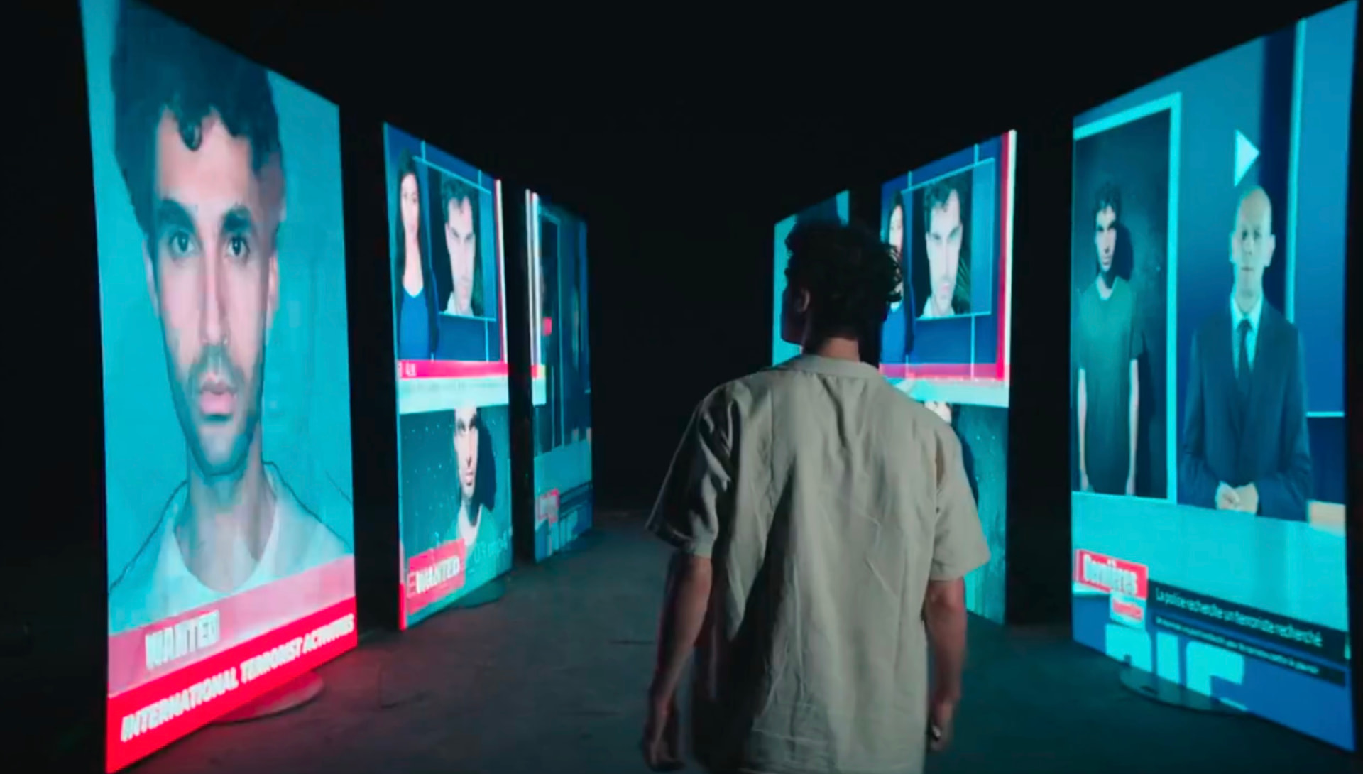 A young man seen from behind walking between rows of glowing screens showing his own wanted news bulletins — اصنع الفرق